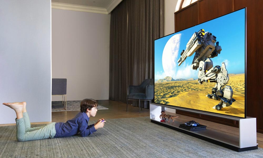 Young boy lying on the floor playing a video game, facing a large flat-screen TV displaying a futuristic desert scene with a giant robotic machine.