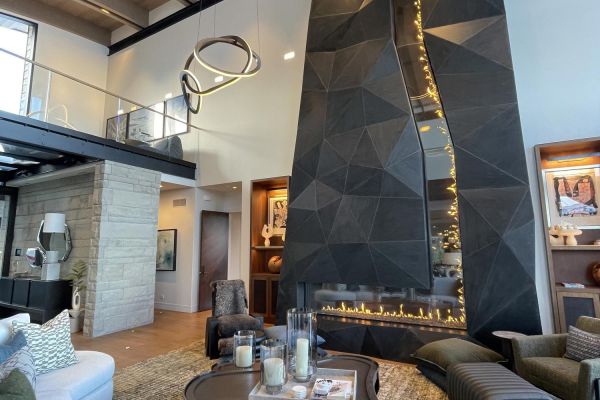 A modern, two-story living area showcasing a striking, geometric black fireplace with built-in lighting, a sleek floating chandelier, and an open mezzanine. Warm wood floors and built-in shelves with decorative items complete the sophisticated space.