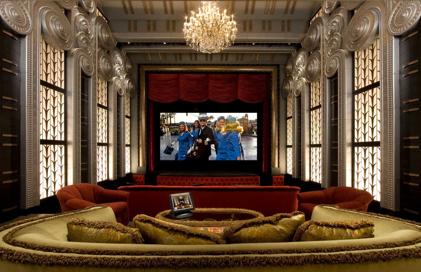 Opulent home theater with curved velvet seating in front of a large screen, flanked by illuminated Art Deco–style wall panels and a central crystal chandelier.