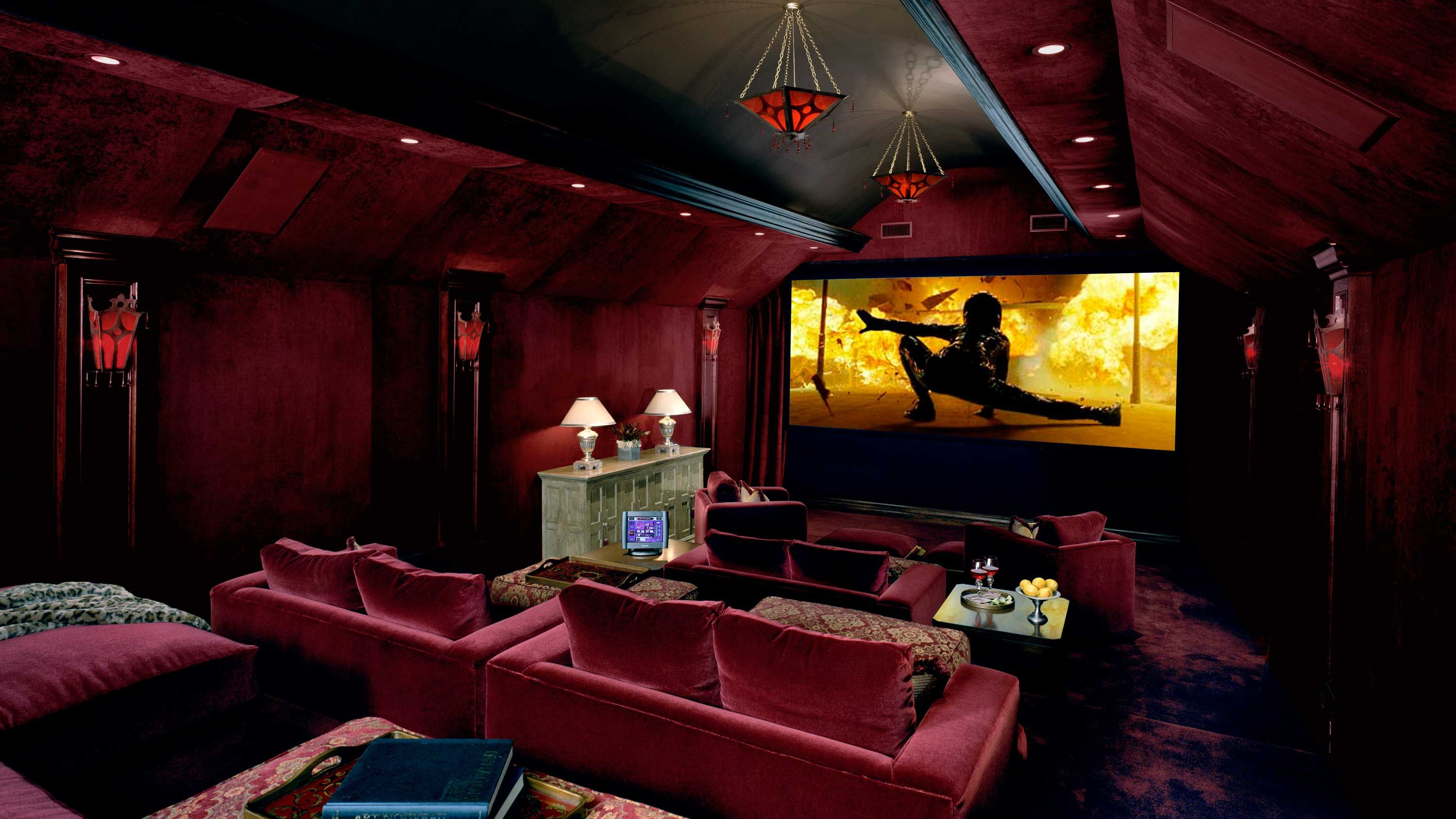 Opulent home theater with deep red walls and plush velvet seating, featuring vintage-style lighting fixtures, a large screen displaying an action scene, and classic décor elements including lamps and a console table.